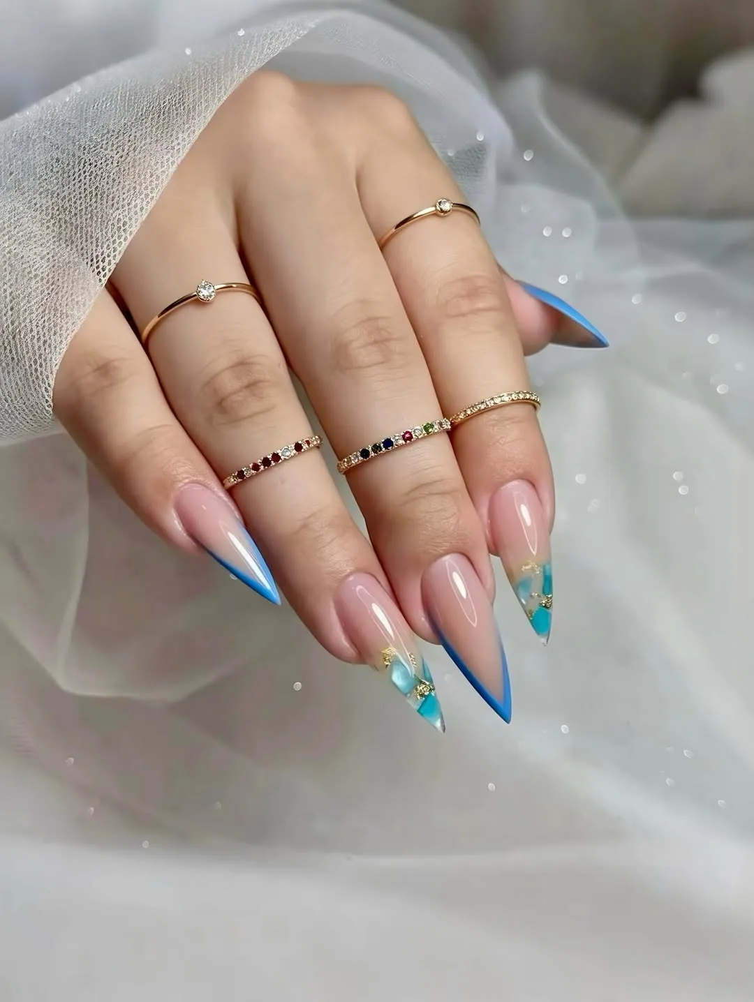 Glossy blue-to-turquoise ombré stiletto nails with aquarium gel-flake accents and gold gem-set rings, over sheer white tulle with floating sparkle
