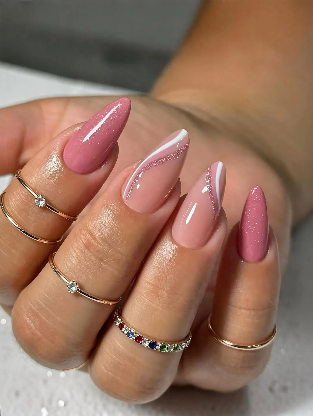 Almond nails in dusty rose and blush with fine white swirl line-art and micro-glitter, hand wearing stacked gold rings on soft grey