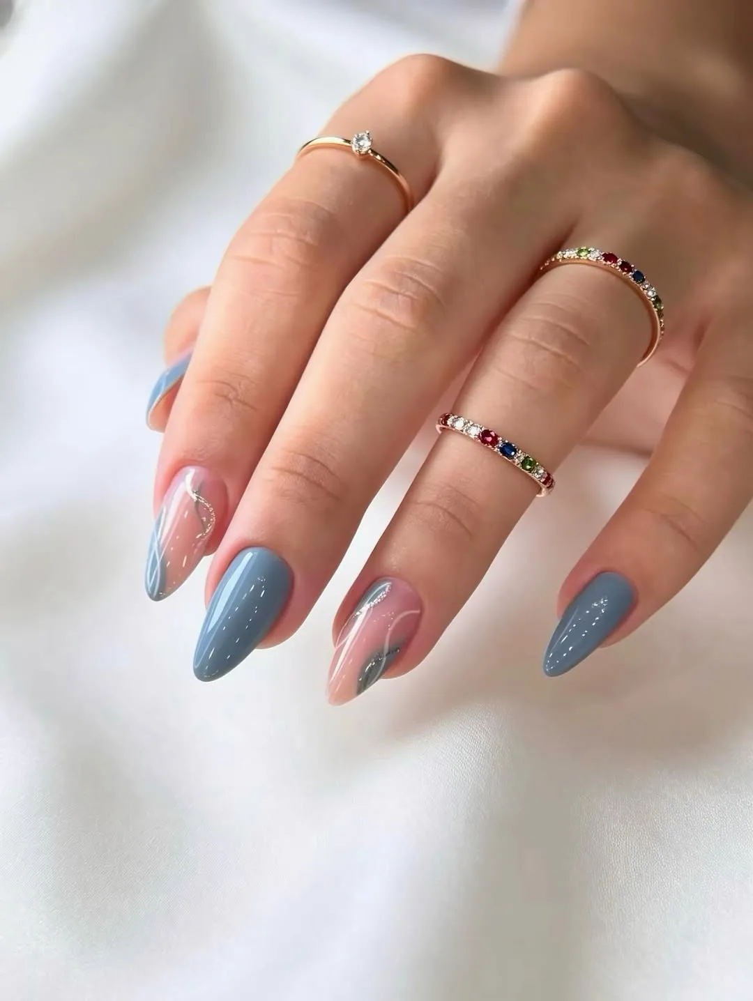 Dusty blue-grey almond nails with clear and silver swirl accents resting on soft white silk with gold jewellery